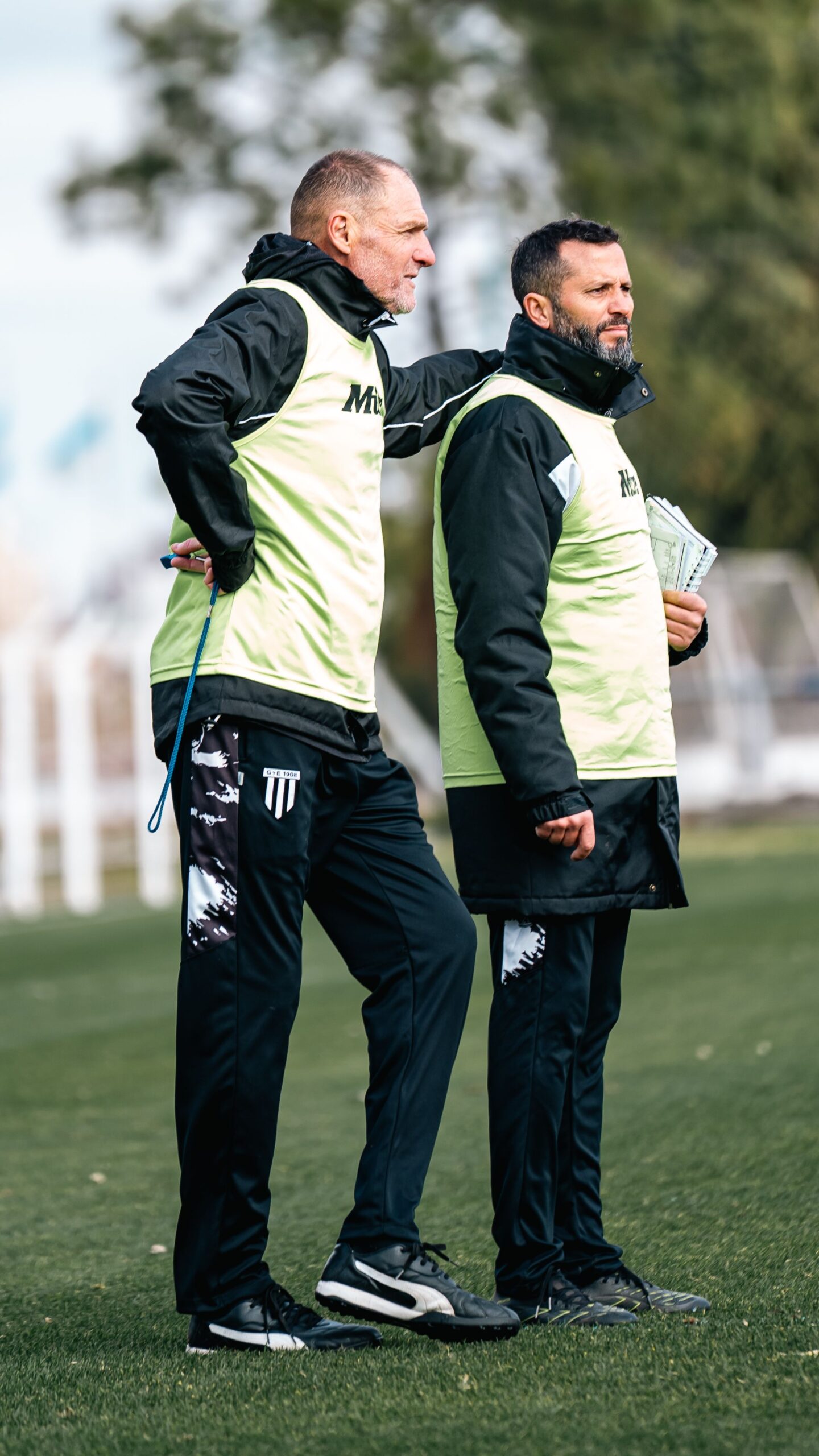 Two soccer coaches in discussion during outdoor training, wearing team vests.